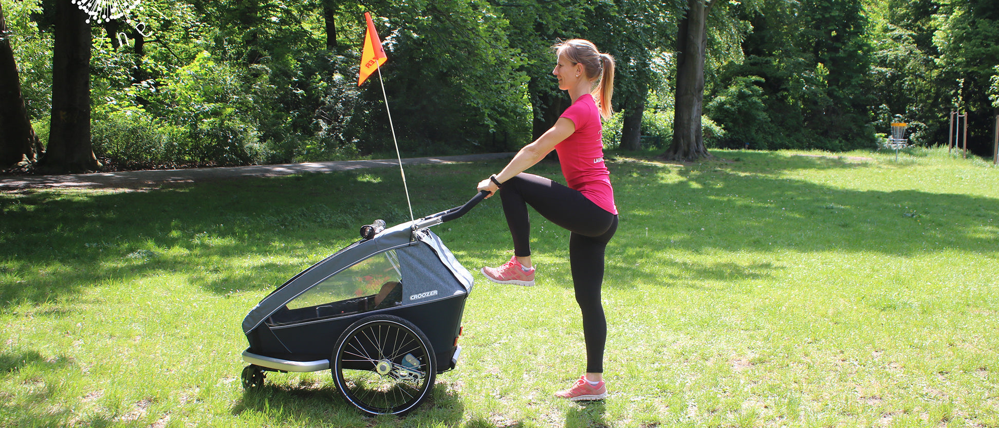 Woman performing standing quad stretch while holding Croozer bike trailer with orange safety flag in park